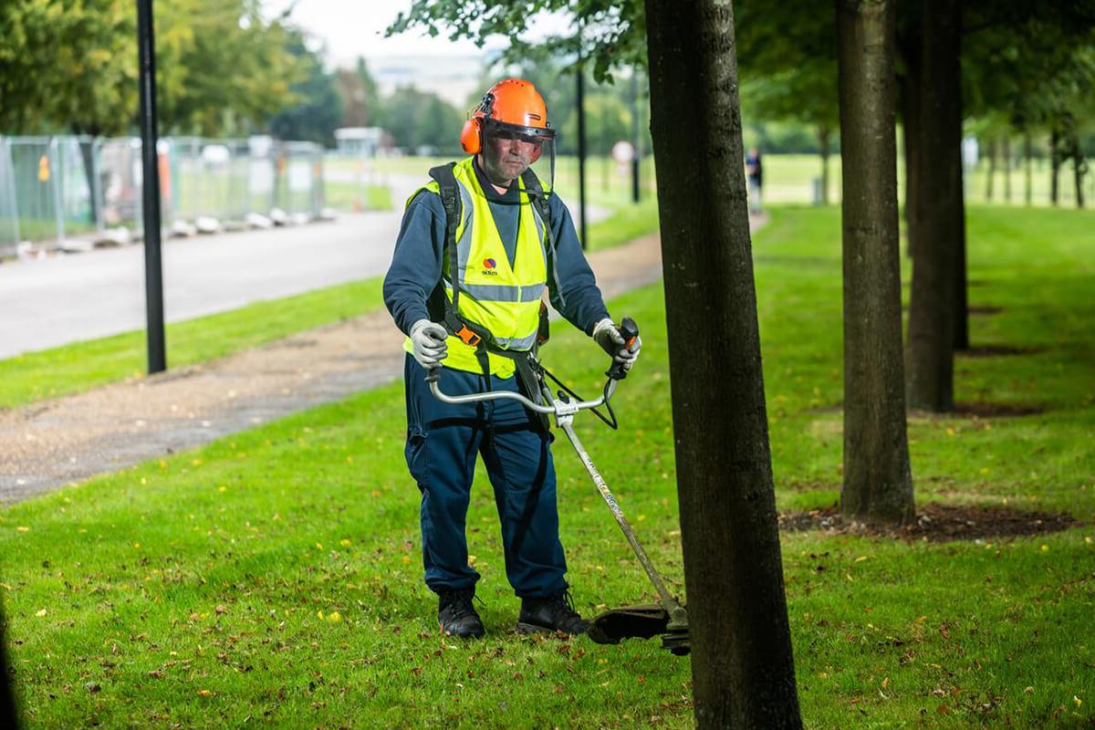 Mitie employee wearing high vis and safety gear using a grass strimmer to conduct ground maintenance